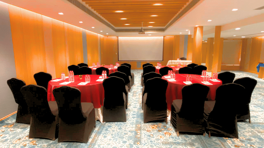 A well-lit conference room at Pride Plaza Ahmedabad, featuring round tables with red tablecloths, chairs, and a projector screen.